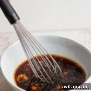 A whisk is stirring a dark-colored sauce in a light gray bowl on a textured surface.