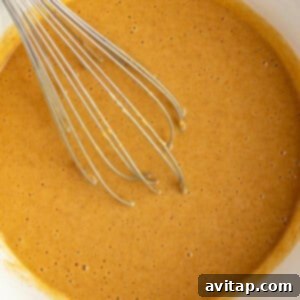 A metal whisk in a white bowl containing smooth, light brown cake batter.