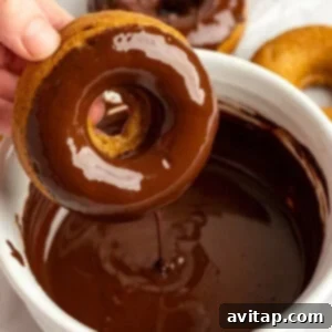 A hand dips a donut into a bowl of chocolate glaze, with more donuts on a white surface in the background.