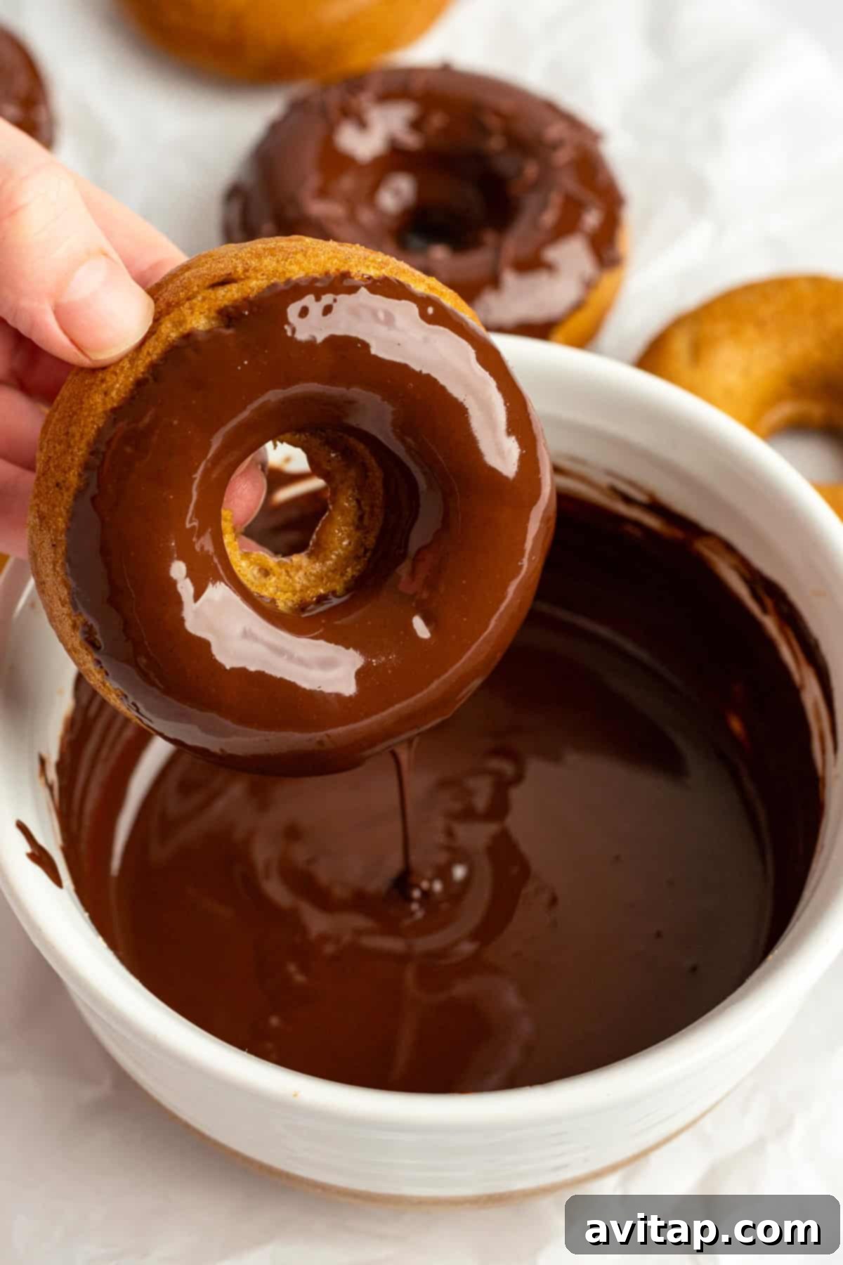 A hand dips a donut into a bowl of chocolate glaze, with more donuts on a white surface in the background.