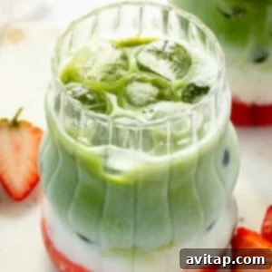 Strawberry Matcha Protein Refresher 11 Layered matcha latte with ice, featuring green matcha, white milk, and red strawberry syrup. Served in a clear, textured glass with sliced strawberries on the side.