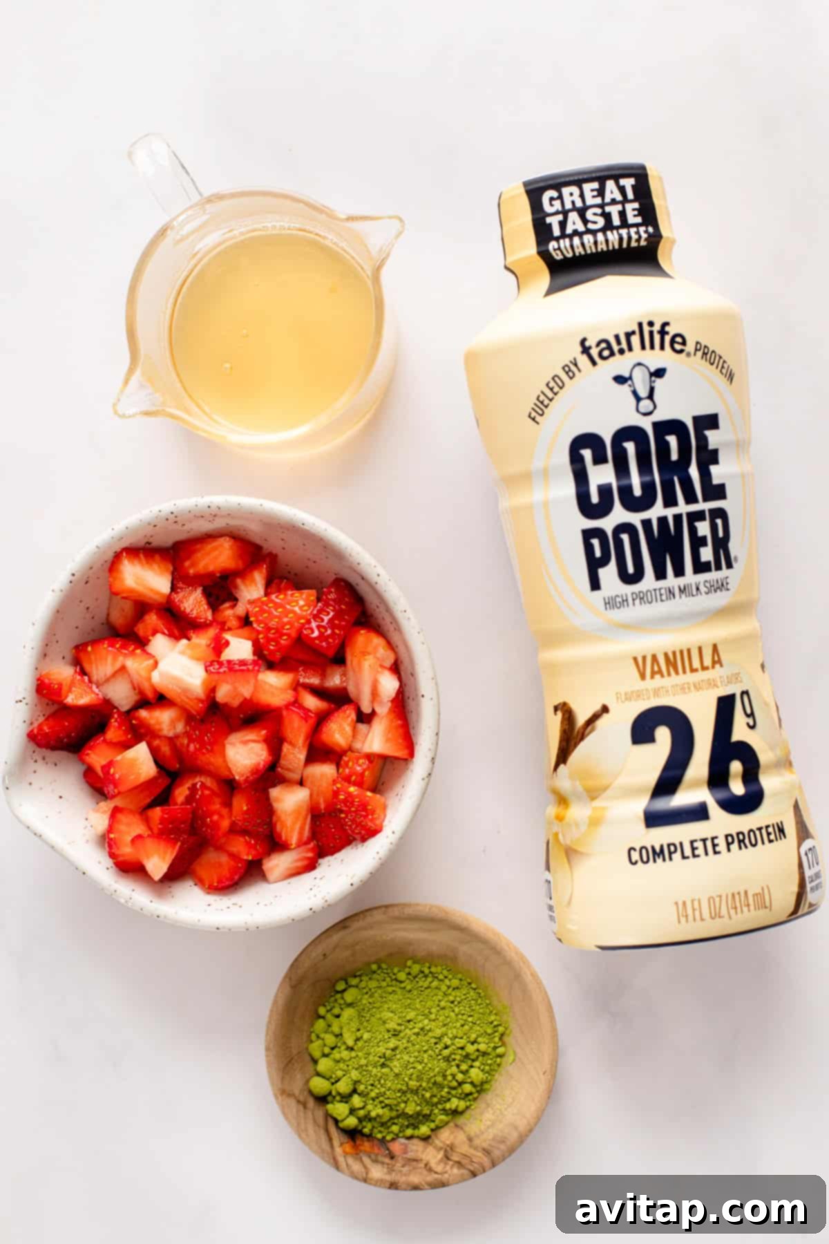 Strawberry Matcha Protein Refresher 3 A selection of key ingredients for the recipe: a bottle of vanilla protein shake, a small jug of honey simple syrup, a bowl of diced fresh strawberries, and a bowl of vibrant green matcha powder, all arranged on a clean white surface.