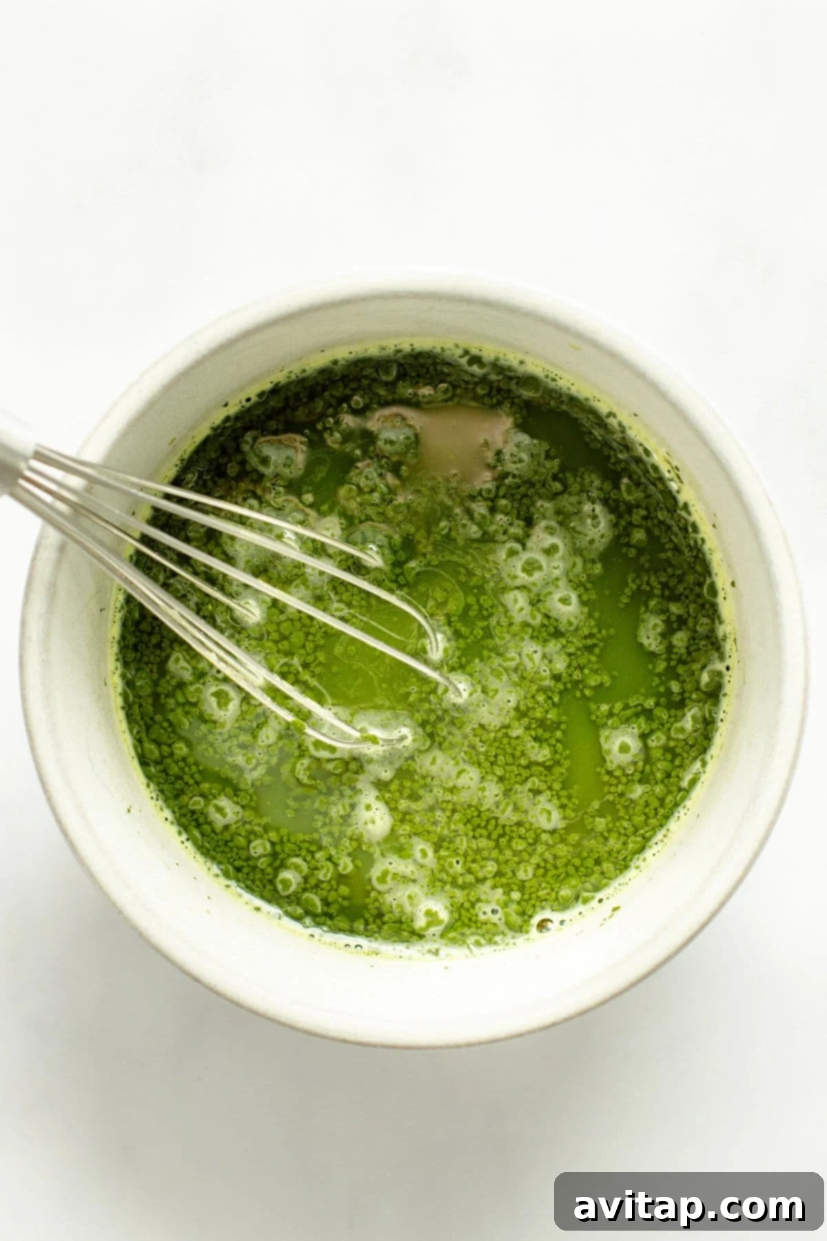 Strawberry Matcha Protein Refresher 4 A traditional bamboo whisk gracefully stirring vibrant green matcha tea in a white bowl, creating a delicate froth against a clean white background.
