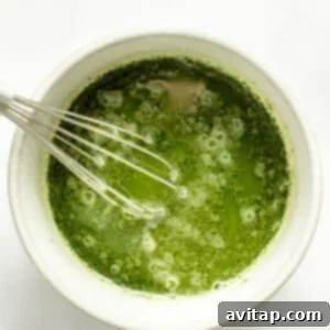 Strawberry Matcha Protein Refresher 8 A whisk in a white bowl containing frothy green matcha tea against a white background.