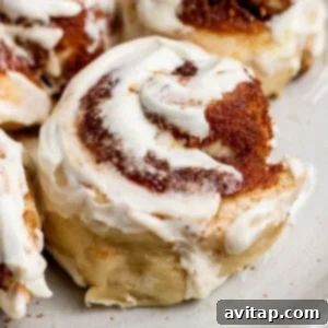 Close-up of a single, freshly baked small batch protein cinnamon roll with creamy white icing and visible cinnamon swirls, resting on a light-colored plate.