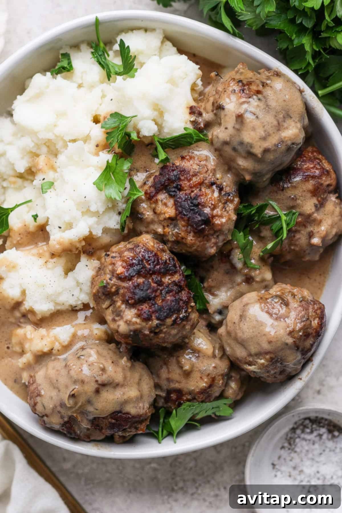 A bowl of meatballs in creamy sauce next to a serving of mashed potatoes. Fresh parsley garnishes the dish.