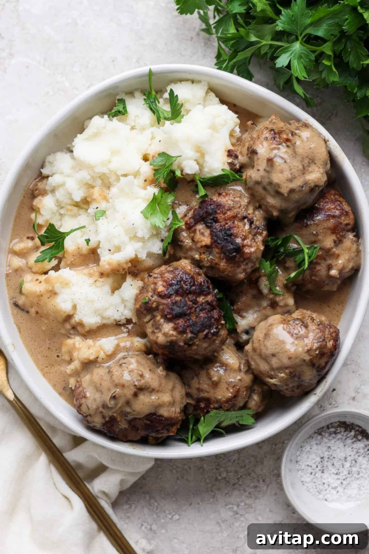 A plate of meatballs covered in gravy, served with mashed potatoes and garnished with fresh parsley. A fork and a small bowl of pepper sit adjacent to the plate.