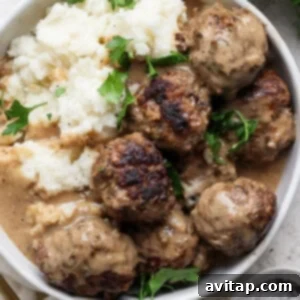 A bowl of meatballs in gravy, served with mashed potatoes, garnished with sprigs of fresh parsley.