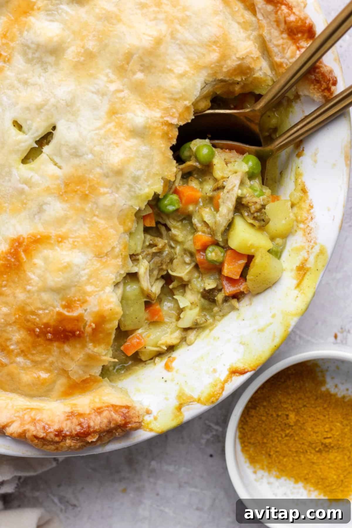 Curried Chicken Pot Pie 2 A pot pie with a flaky crust partially open, revealing a filling of chicken, carrots, peas, and potatoes. Two spoons are placed inside, next to a small dish of curry powder.