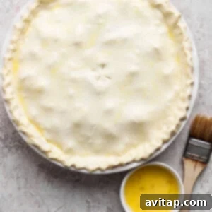 Curried Chicken Pot Pie 9 Unbaked pie with a crimped edge on a round dish, accompanied by a small bowl of egg wash and a pastry brush on a textured surface.