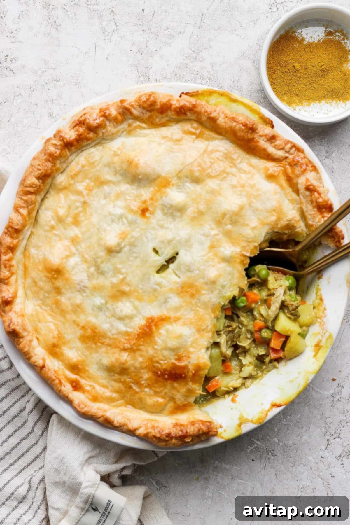Curried Chicken Pot Pie 12 A savory pie with a golden crust is partially sliced, revealing a filling of vegetables and chicken. Two spoons are in the pie, and a bowl of seasoning is nearby.