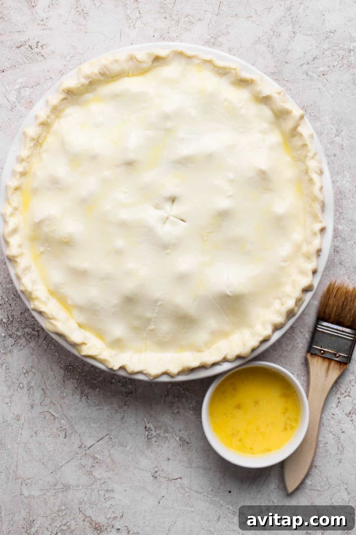 Curried Chicken Pot Pie 3 Unbaked pie with a crimped edge on a round dish, accompanied by a small bowl of egg wash and a pastry brush on a textured surface.