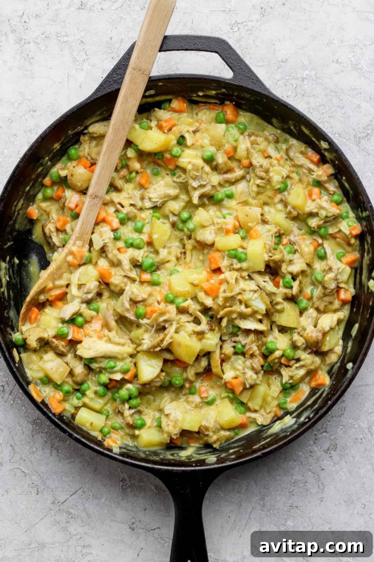 Curried Chicken Pot Pie 4 A skillet of cooked chicken, potatoes, carrots, and peas mixed in a creamy sauce, with a wooden spoon on the side.