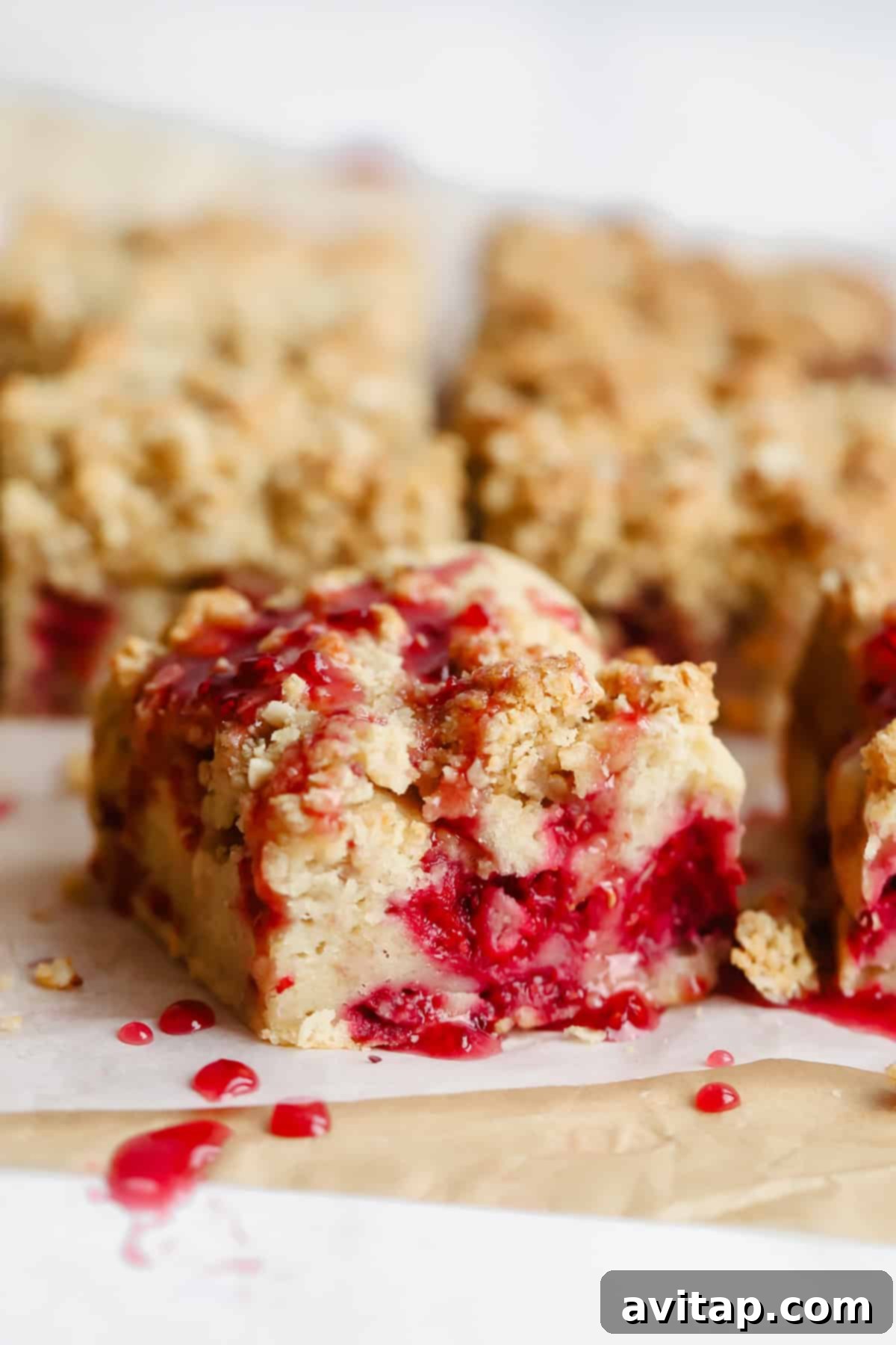 Close-up of golden-brown crumbly dessert bars with a vibrant raspberry filling, topped with a delicate drizzle of red sauce, artfully arranged on parchment paper.