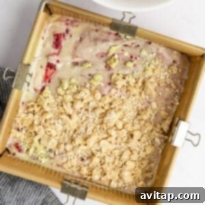 A square baking pan filled with strawberry batter and topped with a streusel crumble, positioned next to a bowl of additional crumble on a white surface with a dark striped cloth.