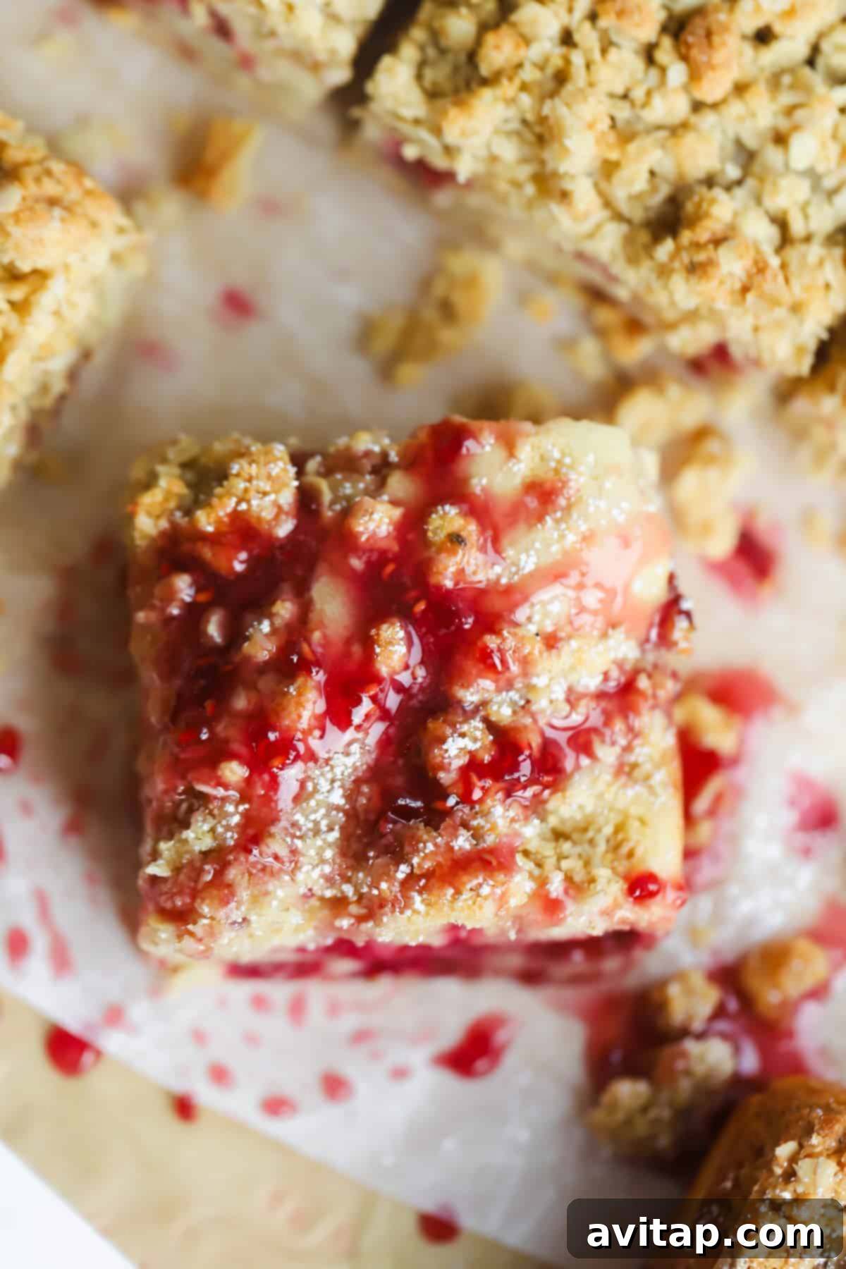 A single square piece of dessert, generously topped with a crumbly streusel and elegantly drizzled with a vibrant red fruit sauce, presented on parchment paper.