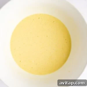 A bowl containing smooth, yellow batter, slightly frothy, resting on a clean white surface, indicating wet ingredients have been mixed.