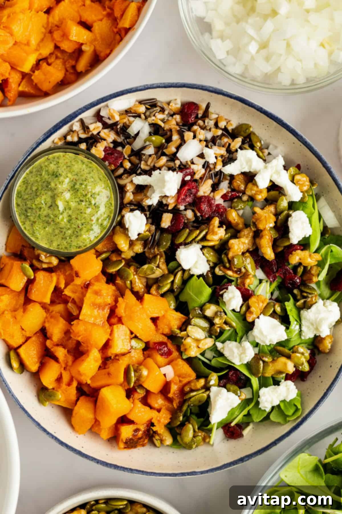 A vibrant salad bowl featuring mixed greens, caramelized roasted butternut squash, crumbled goat cheese, crunchy candied nuts, and a blend of grains, accompanied by a small side of bright green tarragon dressing.