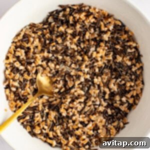 A white serving bowl filled with a perfectly mixed blend of cooked wild rice and farro, highlighting the textures and colors of the grains. A gold spoon is nestled within the grains.