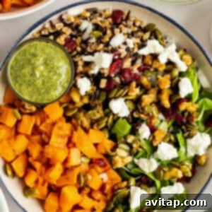 A beautifully assembled Hodgepodge Butternut Squash Bowl, featuring a colorful array of mixed greens, roasted squash, goat cheese, candied nuts, grains, and dried cranberries, served alongside a small container of vibrant green dressing.