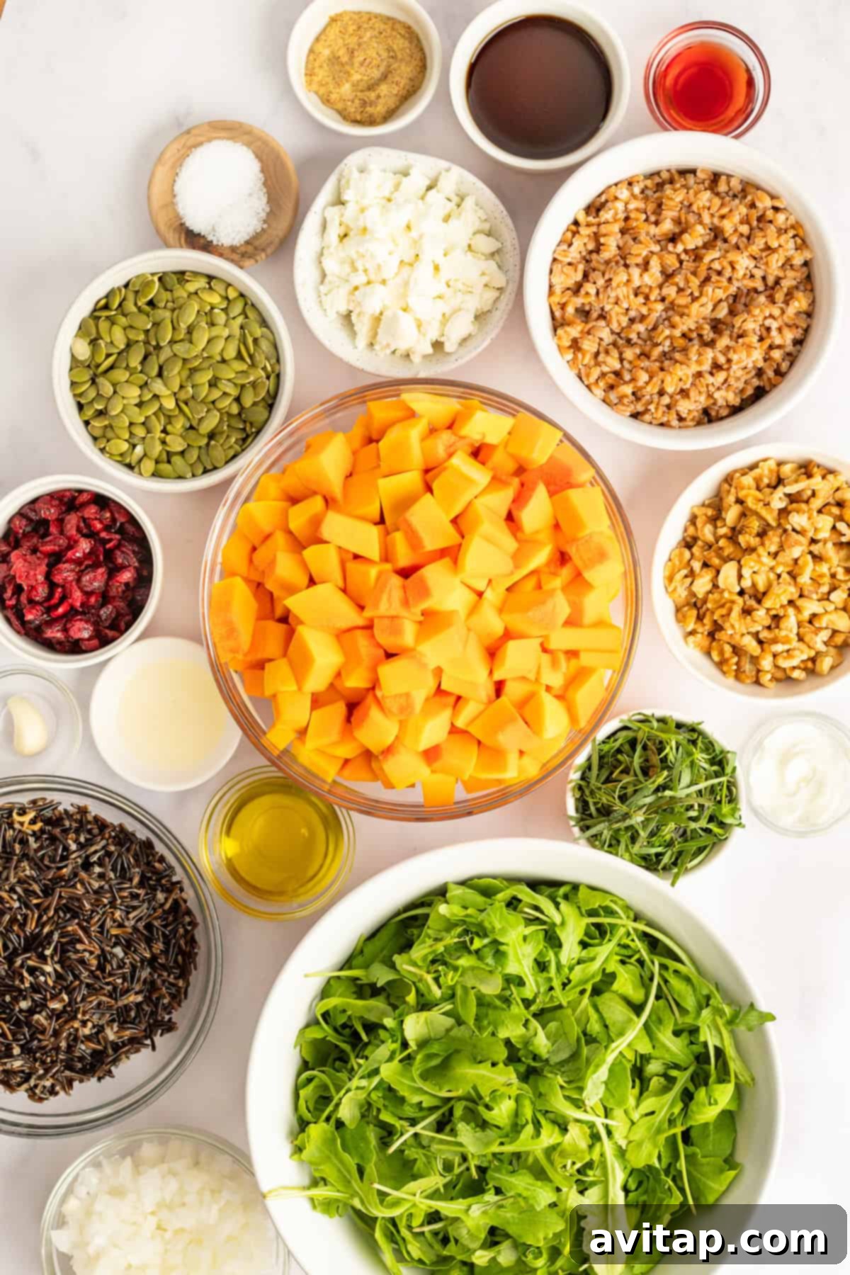 An overhead view of various fresh ingredients laid out on a wooden table, including bright green arugula, uniformly cubed butternut squash, a mix of walnuts and pepitas, a bowl of cooked grains, crumbled goat cheese, and small dishes of liquids for the dressing.