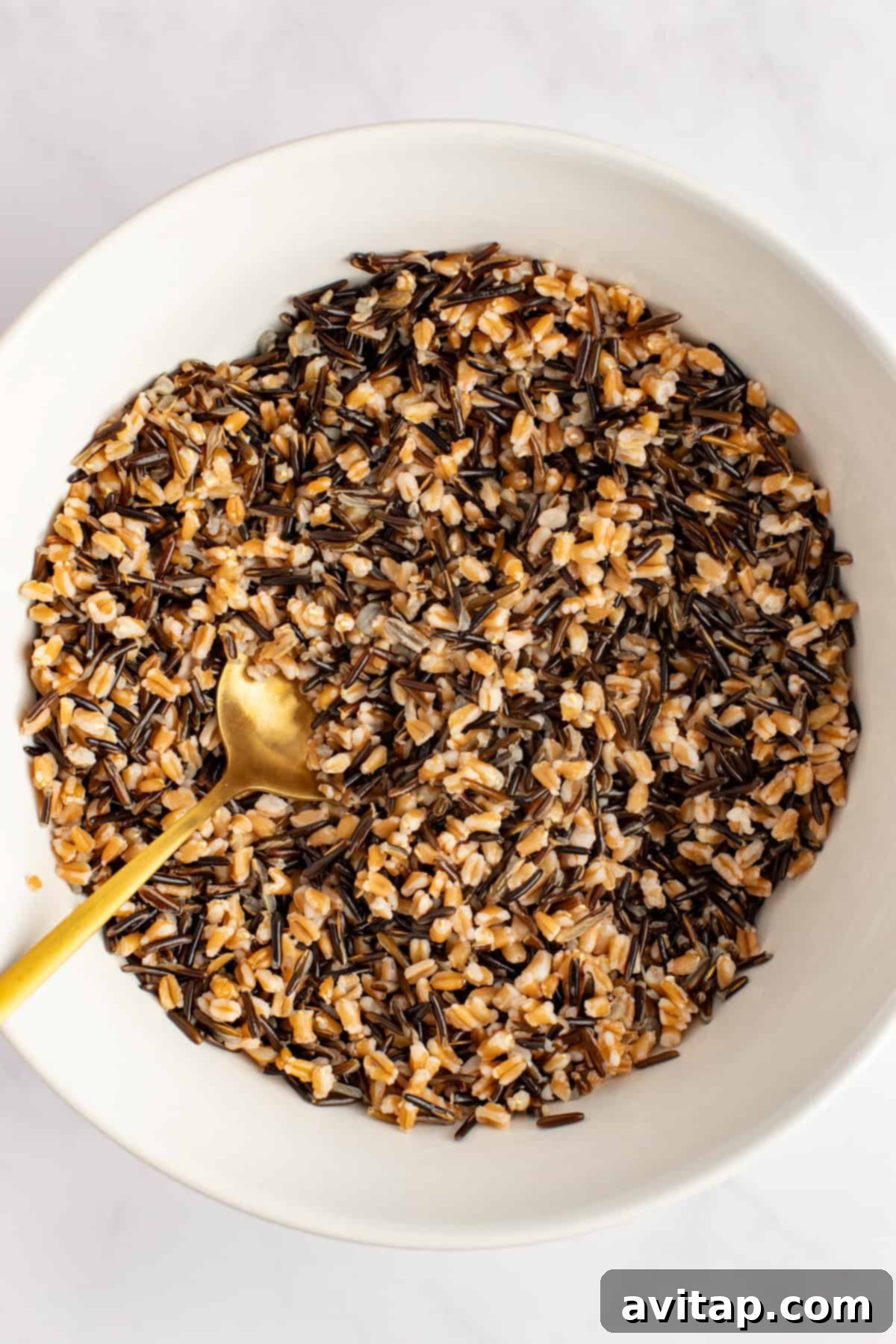 A beautifully composed close-up of a white bowl filled with a harmonious blend of cooked wild rice and farro, showcasing their distinct textures and colors. A gold-toned spoon rests gently in the grains.
