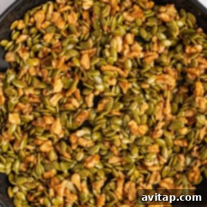 A pan filled with a mix of walnuts and green pepitas, coated in a sticky maple syrup glaze, actively toasting on a stovetop.