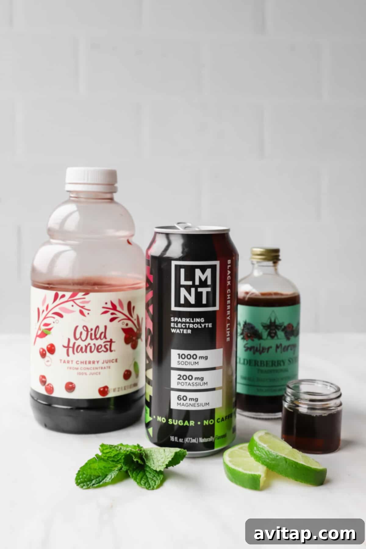 A bottle of tart cherry juice, a can of LMNT electrolyte drink, elderberry syrup, mint leaves, and lime slices on a white surface.