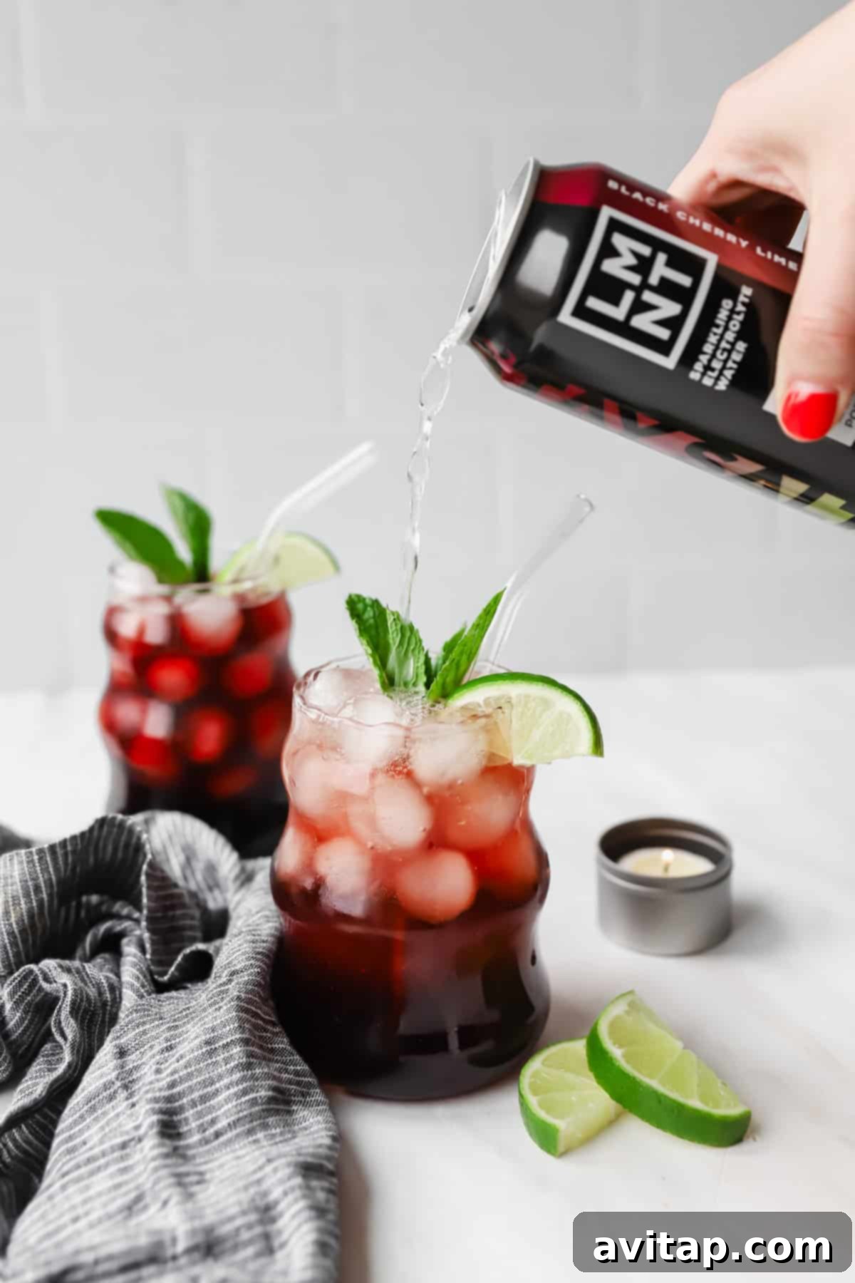 A hand pours a black cherry drink into a glass filled with ice, cherries, lime slices, and mint. Another similar drink is in the background, next to lime wedges and a small candle.