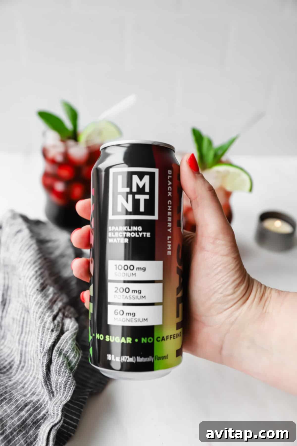 Hand holding a can of LMNT black cherry lime sparkling electrolyte water. Two glasses with red drinks and lime wedges are in the background.