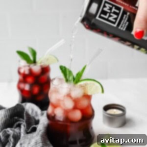 A hand pours a black cherry drink into a glass filled with ice, cherries, lime slices, and mint. Another similar drink is in the background, next to lime wedges and a small candle.