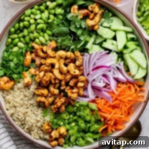A vibrant, colorful salad arranged in a large bowl, featuring a mix of cashews, edamame, quinoa, sliced cucumbers, shredded carrots, red onions, and various fresh herbs. Adjacent to it are small bowls holding creamy dressing and additional chopped carrots.