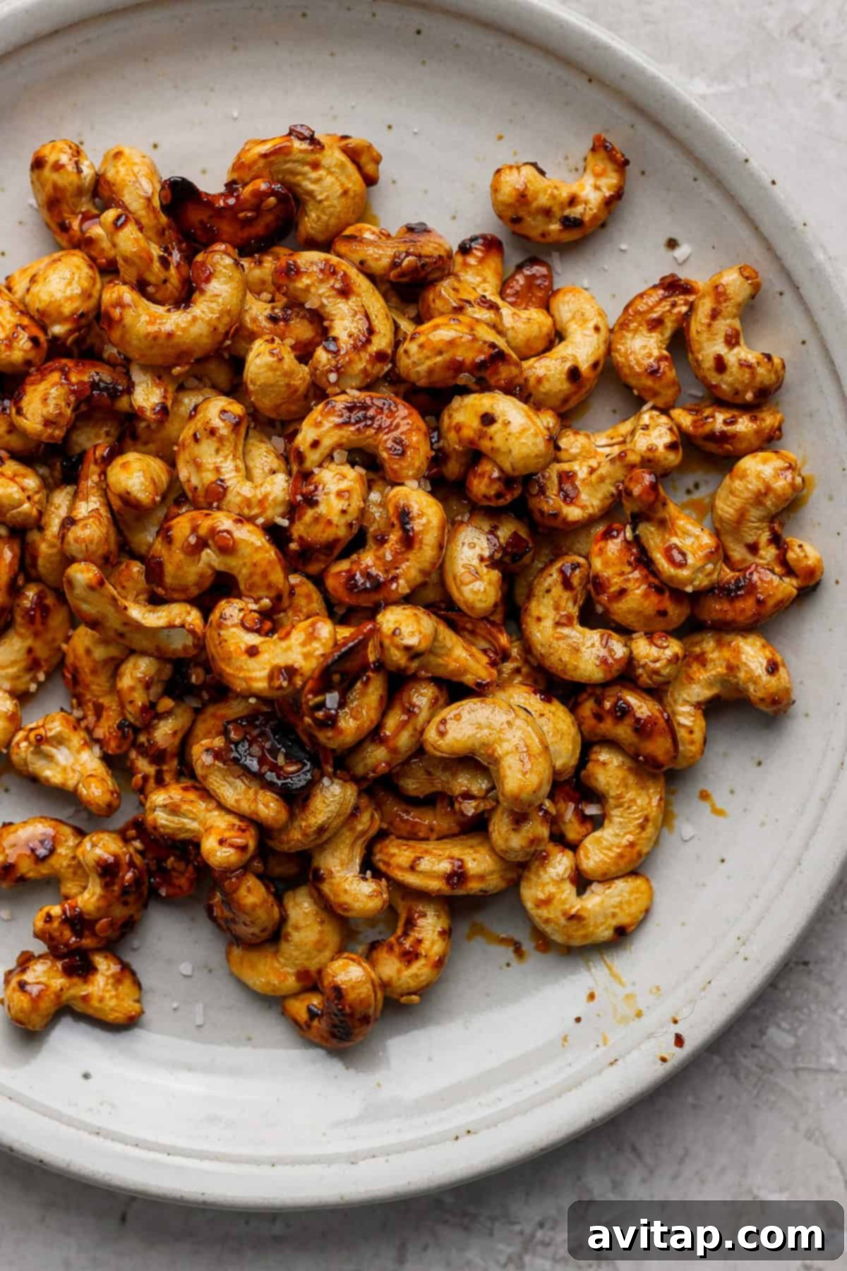 A close-up shot of perfectly roasted cashews, glistening with a chili flake and salt seasoning, presented on a minimalist light gray surface.