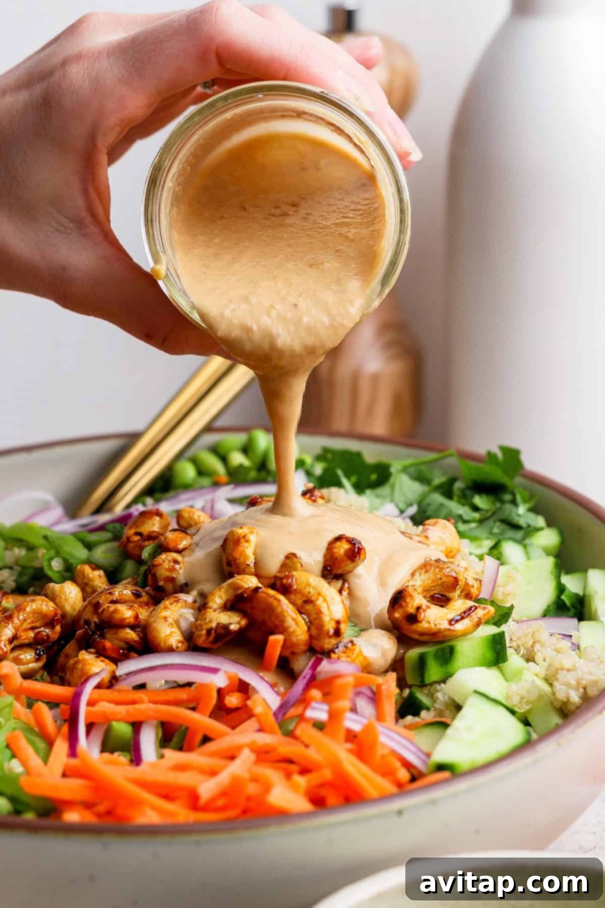 A hand gently drizzles rich, creamy dressing over a vibrant salad featuring shredded carrots, cucumber slices, red onions, and an array of fresh greens in a rustic bowl.