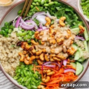 A bowl of vibrant salad featuring quinoa, edamame, sliced red onions, cucumbers, carrots, crunchy cashews, and fresh green onions, generously topped with a creamy dressing.