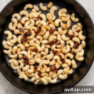 A black skillet containing roasted cashews, visibly coated with a spicy seasoning blend, set against a dark, textured background.