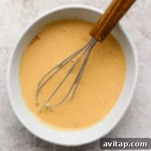 A whisk poised within a white bowl, stirring a pale orange mixture, resting on a subtly speckled light-colored countertop.