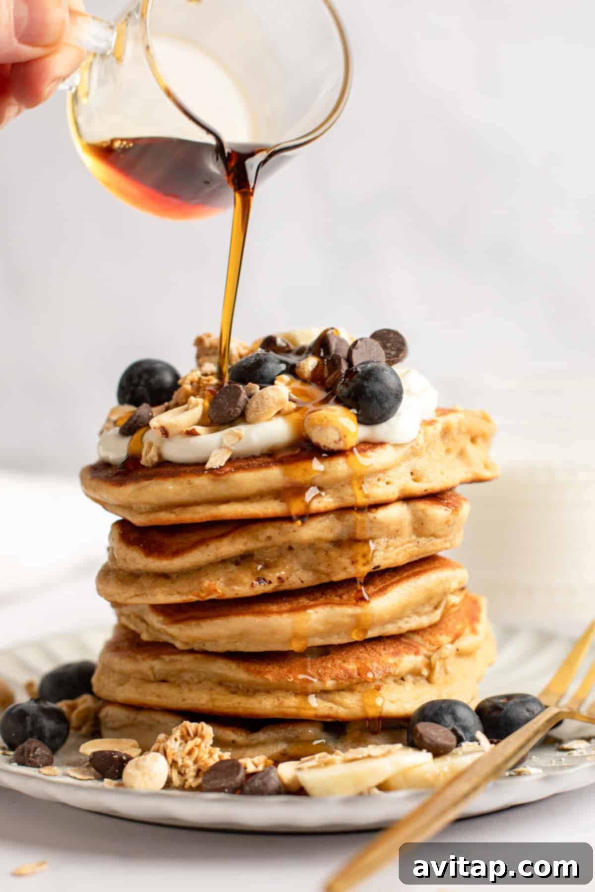 A beautifully stacked tower of golden-brown protein pancakes, generously adorned with fresh blueberries, rich chocolate chips, crunchy granola, and a sprinkle of nuts. A stream of maple syrup cascades over the stack, inviting indulgence.