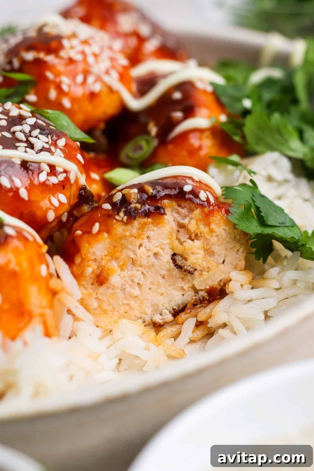 Sticky Sesame Ginger Salmon Meatballs 7 Close-up of rice topped with saucy salmon meatballs, garnished with sesame seeds and cilantro. A meatball is cut open, revealing its interior. Drizzled sauce is visible on top.