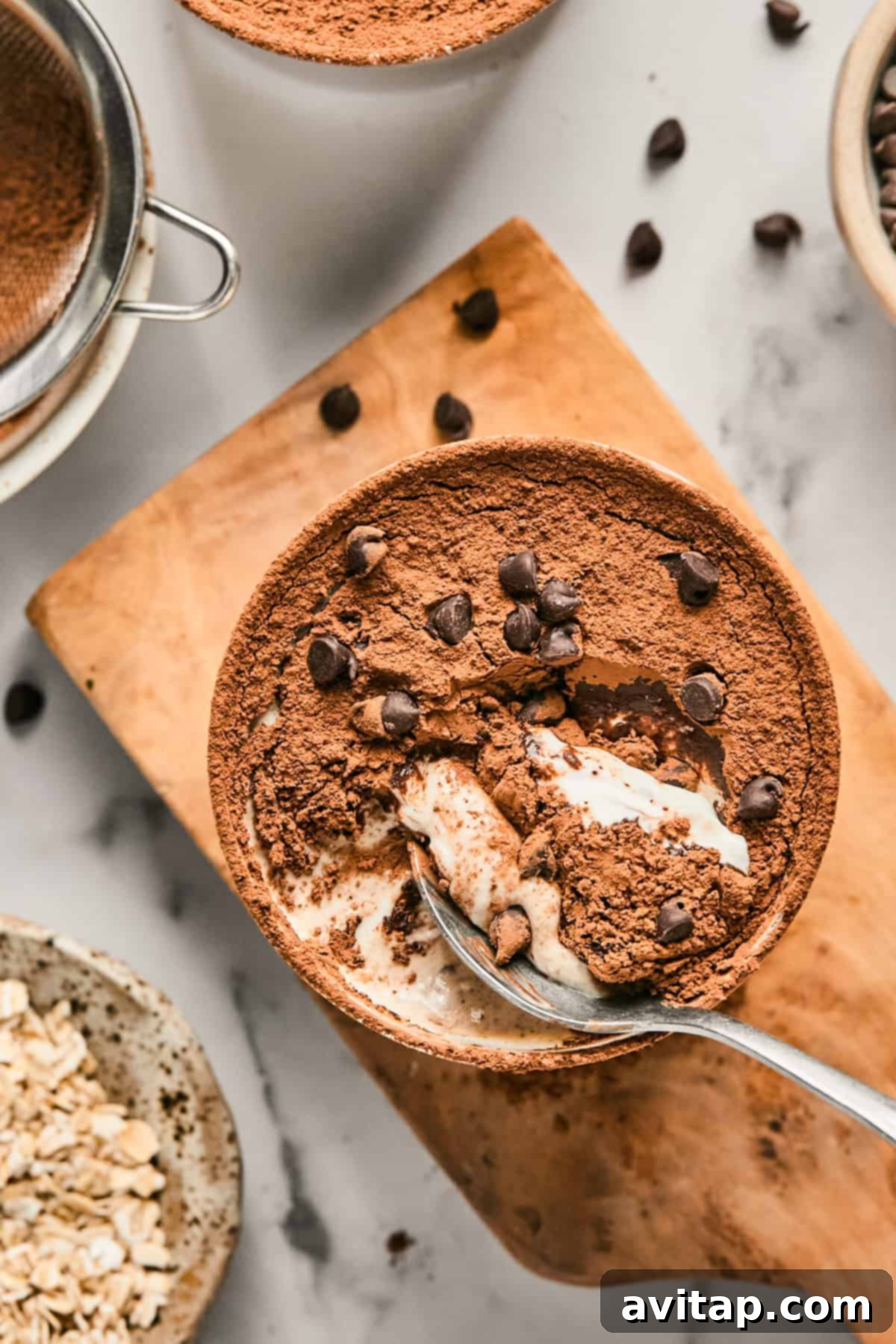 Bowl of Tiramisu Blended Oats topped with cocoa powder and chocolate chips on a wooden board, next to a sieve, oats, and scattered chocolate chips. A spoon is scooping a portion from the bowl.
