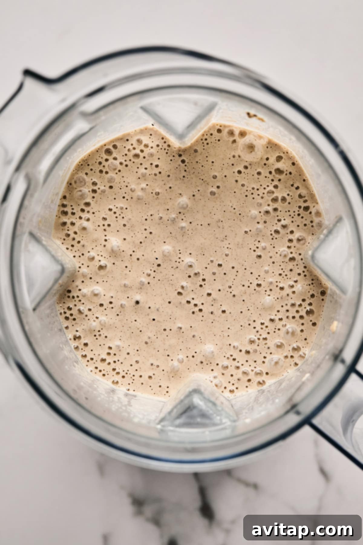Top view of a blender filled with a frothy beige liquid, freshly blended oats, on a white marble surface.