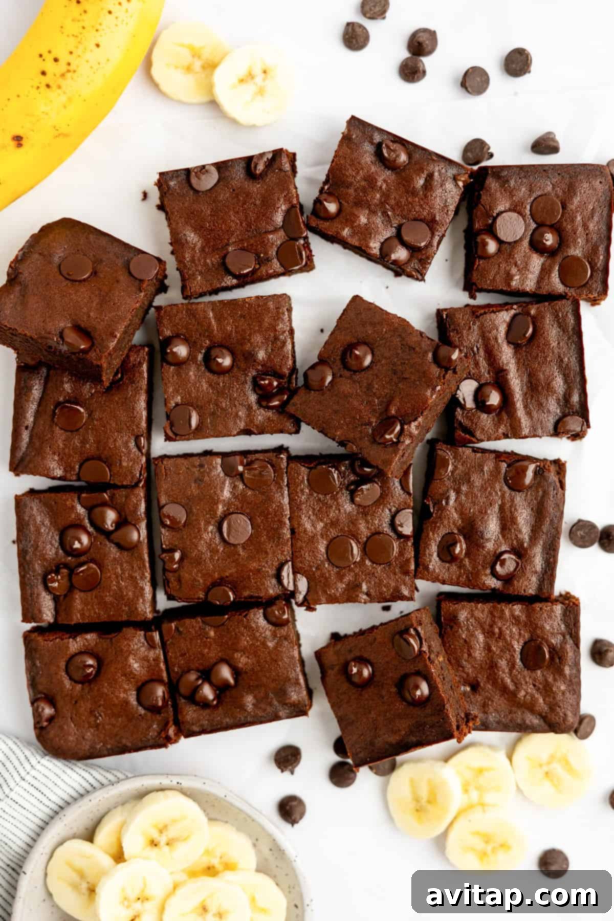 Banana Bread Brownie Mashup 2 Brownies with chocolate chips arranged on a surface, surrounded by banana slices and a whole banana.