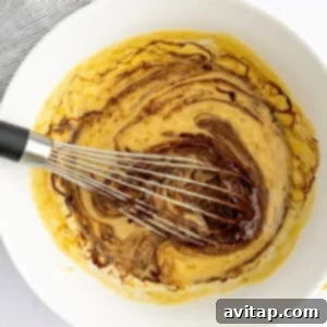 Banana Bread Brownie Mashup 10 A large white bowl with a whisk, mixing ingredients that include a swirled liquid and batter, next to a whole banana on a white surface.