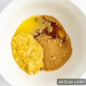 Banana Bread Brownie Mashup 8 A white bowl containing mashed bananas, brown sugar, and melted butter on a white surface.