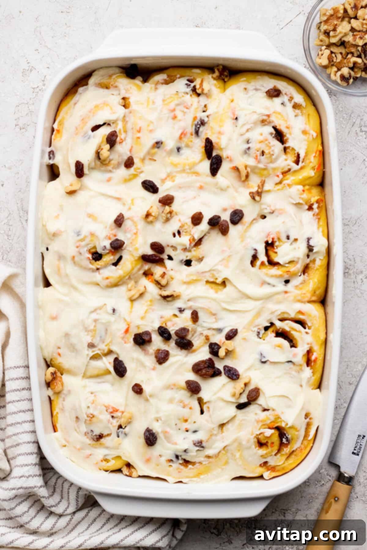 Carrot Cake Cinnamon Swirls 2 A baking dish filled with frosted carrot cake cinnamon rolls topped with raisins and walnuts. A knife and striped cloth are nearby, suggesting a delightful breakfast scene.