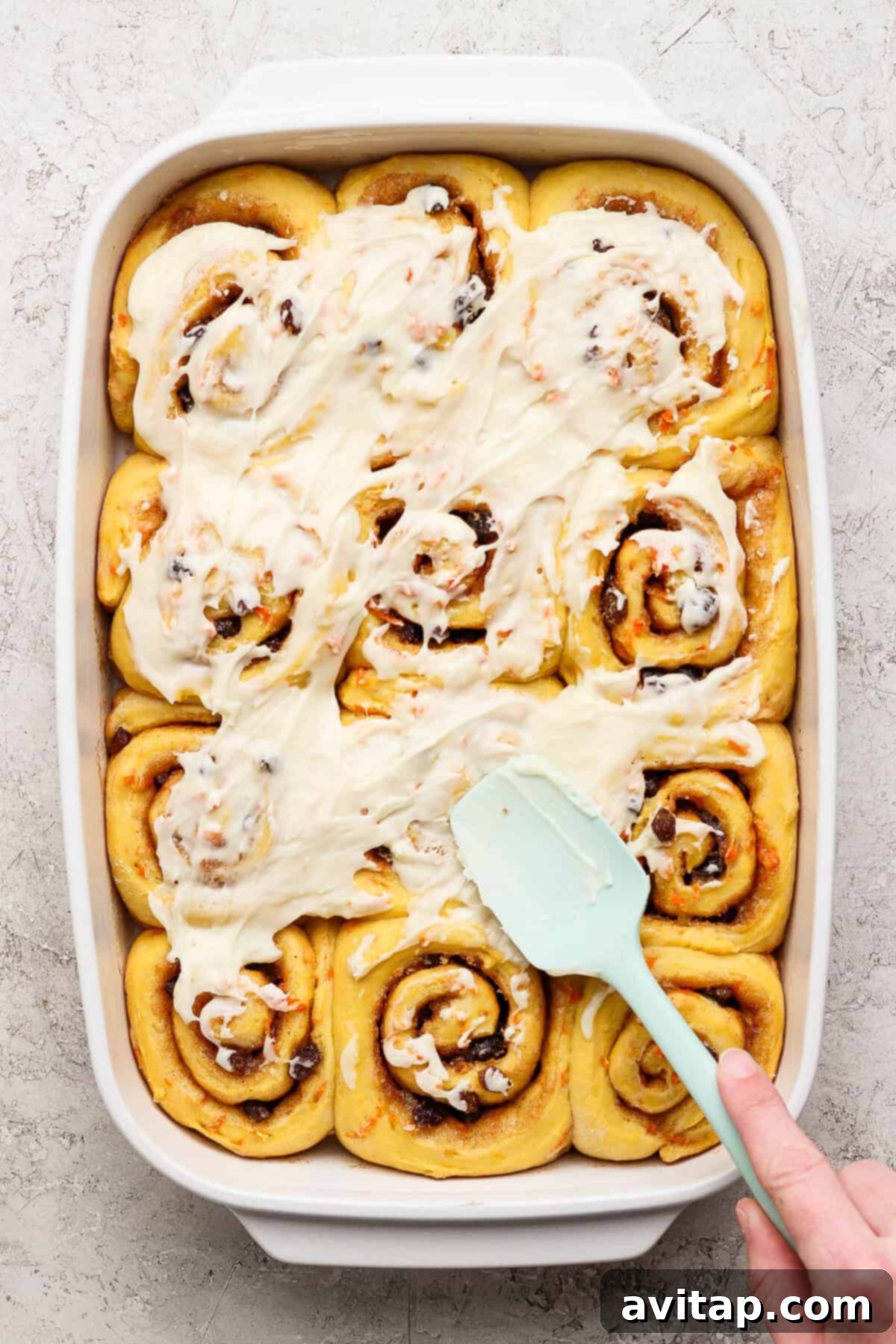 Carrot Cake Cinnamon Swirls 5 A person spreads white cream cheese icing over freshly baked carrot cake cinnamon rolls arranged neatly in a rectangular baking dish, ready for serving.
