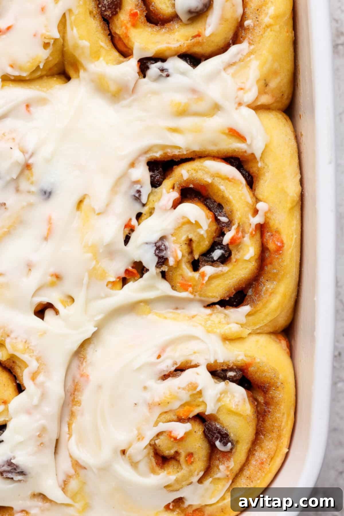 Carrot Cake Cinnamon Swirls 6 Close-up of frosted carrot cake cinnamon rolls in a baking dish, featuring swirls of dough with cinnamon and raisin filling, topped with luscious cream cheese icing, highlighting the tempting texture.