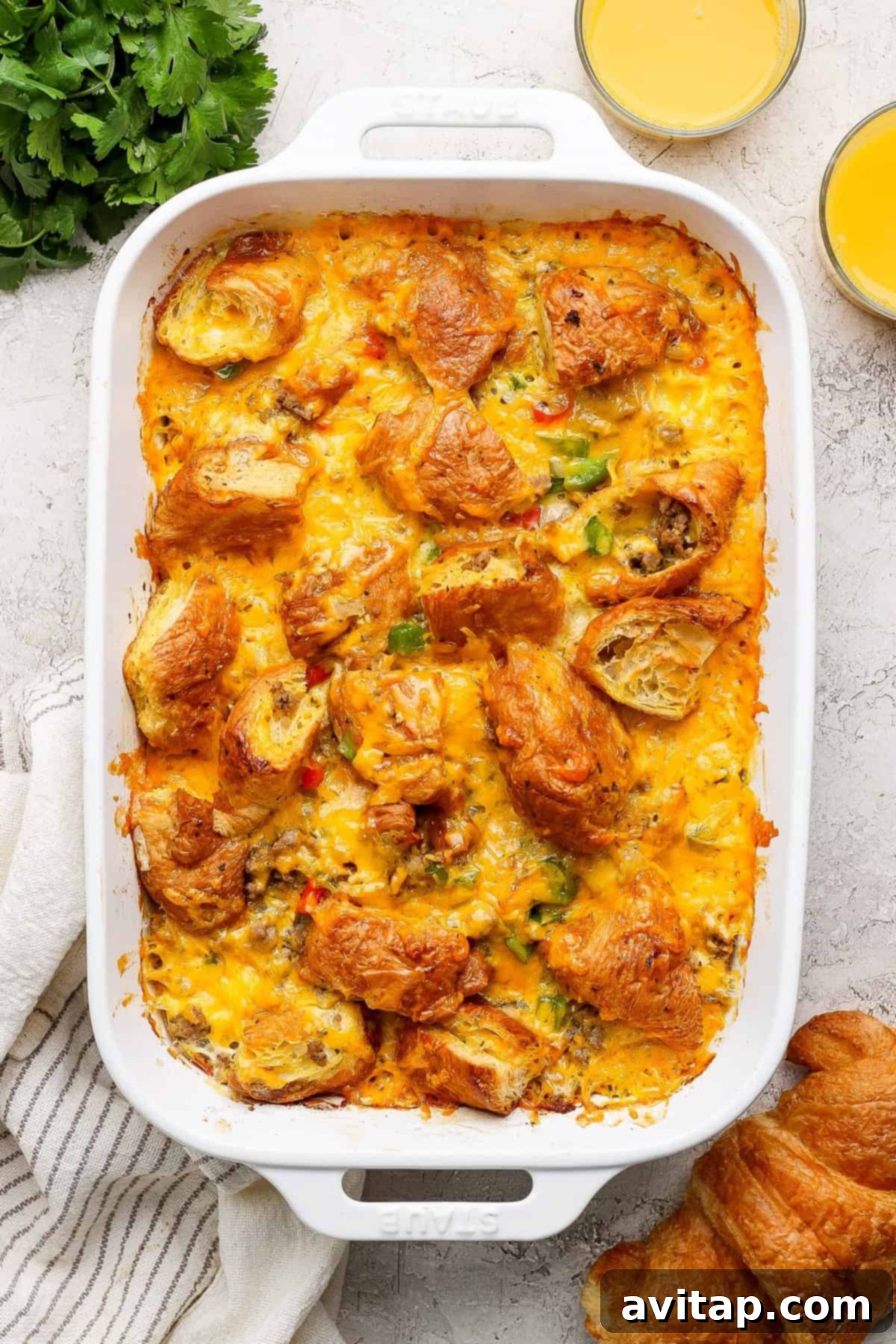 A golden-brown baked croissant egg bake in a white casserole dish, garnished with fresh herbs. Two glasses of orange juice sit beside it, ready for a delightful breakfast or brunch.