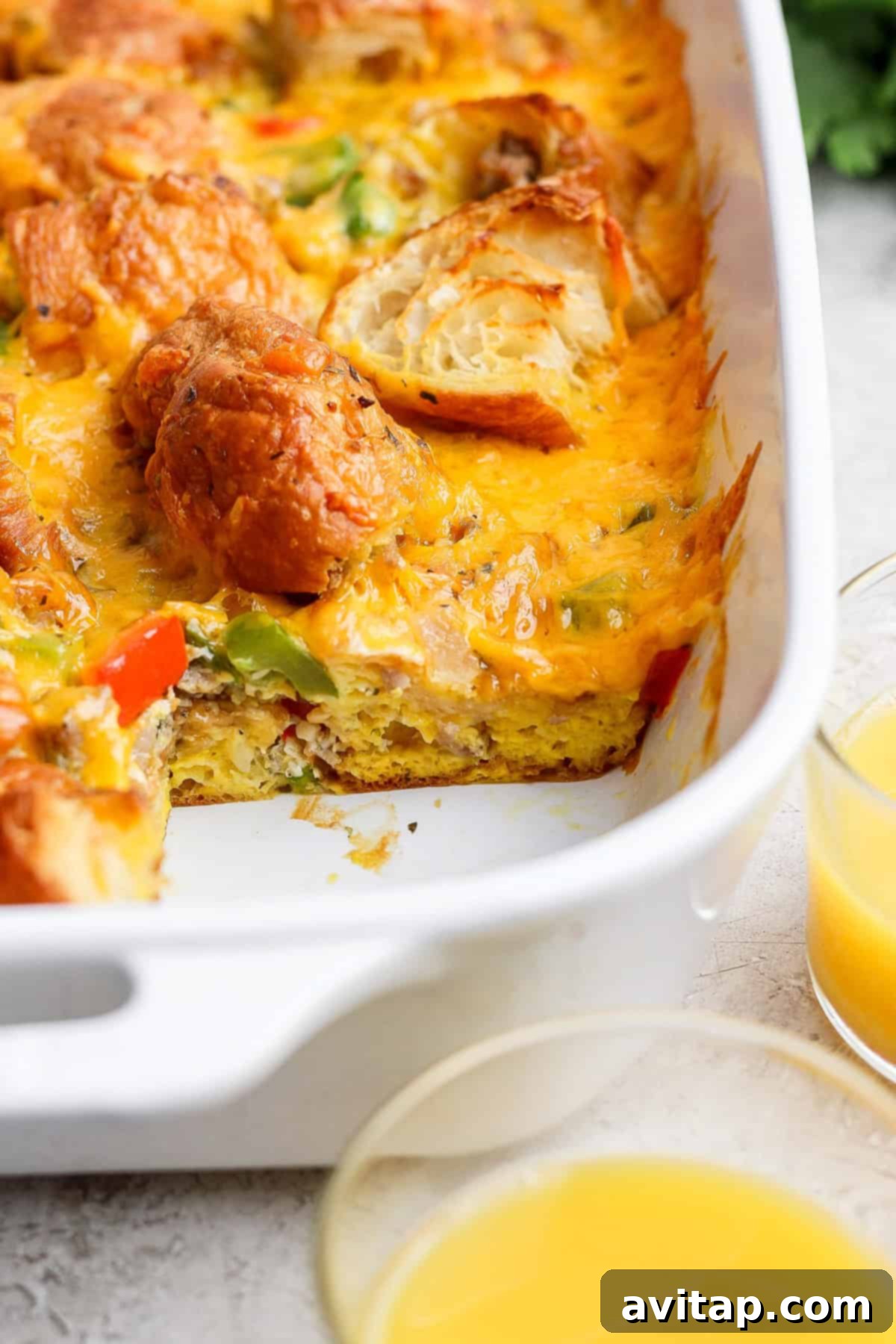 A delicious breakfast casserole with melted cheese, vegetables, and croissant pieces, perfectly baked in a white dish, served with orange juice.