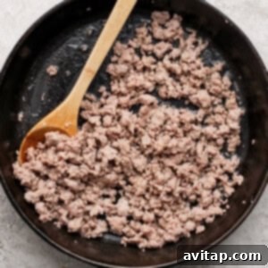 Ground breakfast sausage cooking in a black cast-iron skillet, being broken up with a wooden spoon.